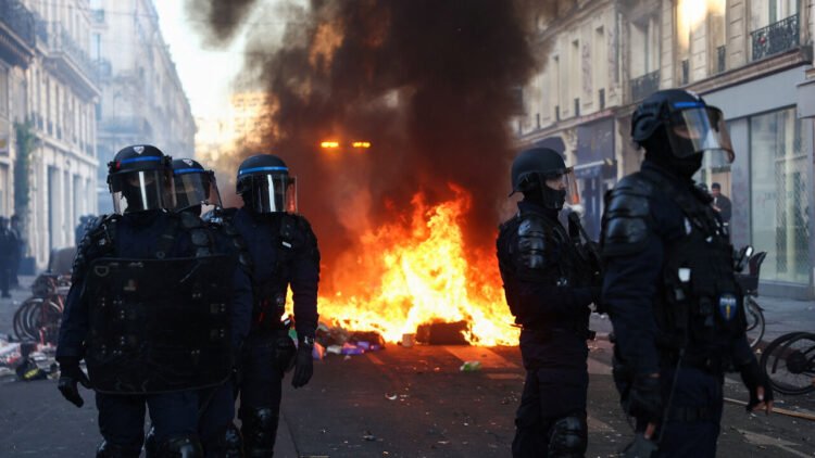 A fire burns, as Police officers in riot gear stand guard, during civil unrest, the day after a national day of demonstrations against the cost of living and rising fuel prices, in Paris, France.